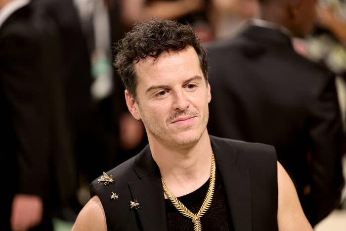 Andrew Scott in a dark outfit adorned with insect brooches, and a gold chain necklace at a formal event