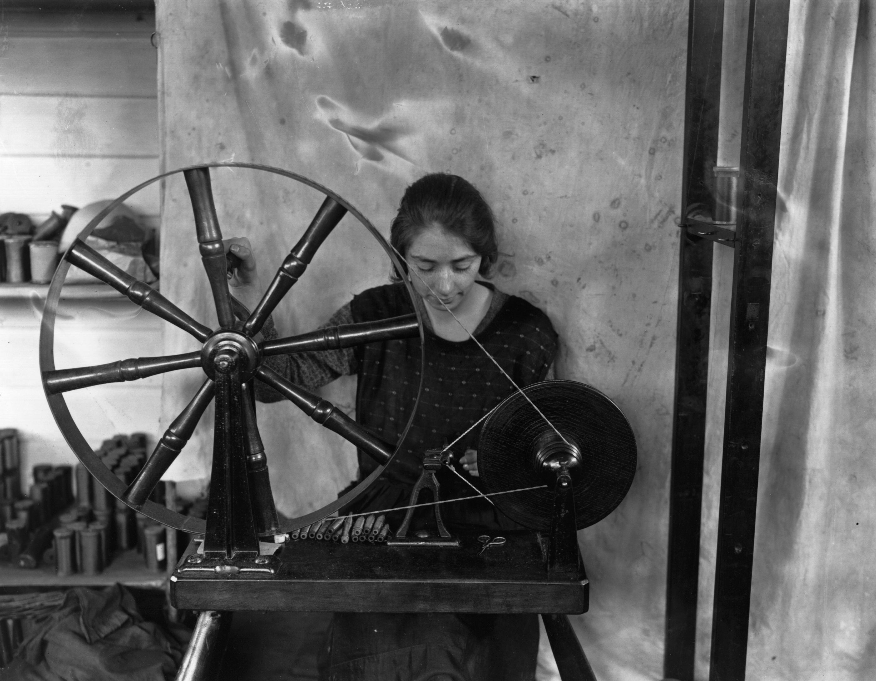 A woman operates a spinning wheel for weaving. She focuses on her work with various tools and materials in the background