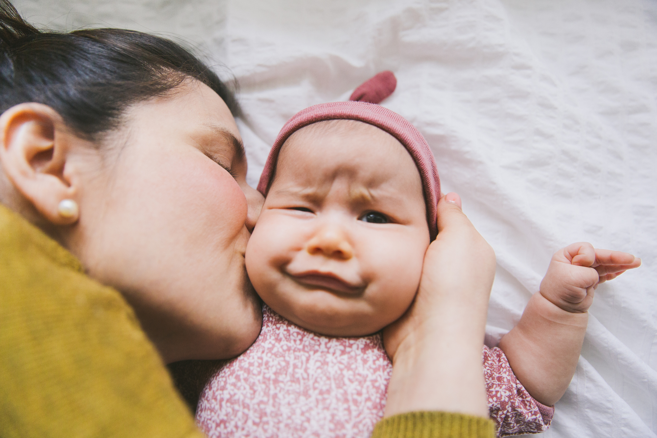 A woman kisses a baby on the cheek while the baby makes a funny face. The baby wears a pink hat and patterned onesie