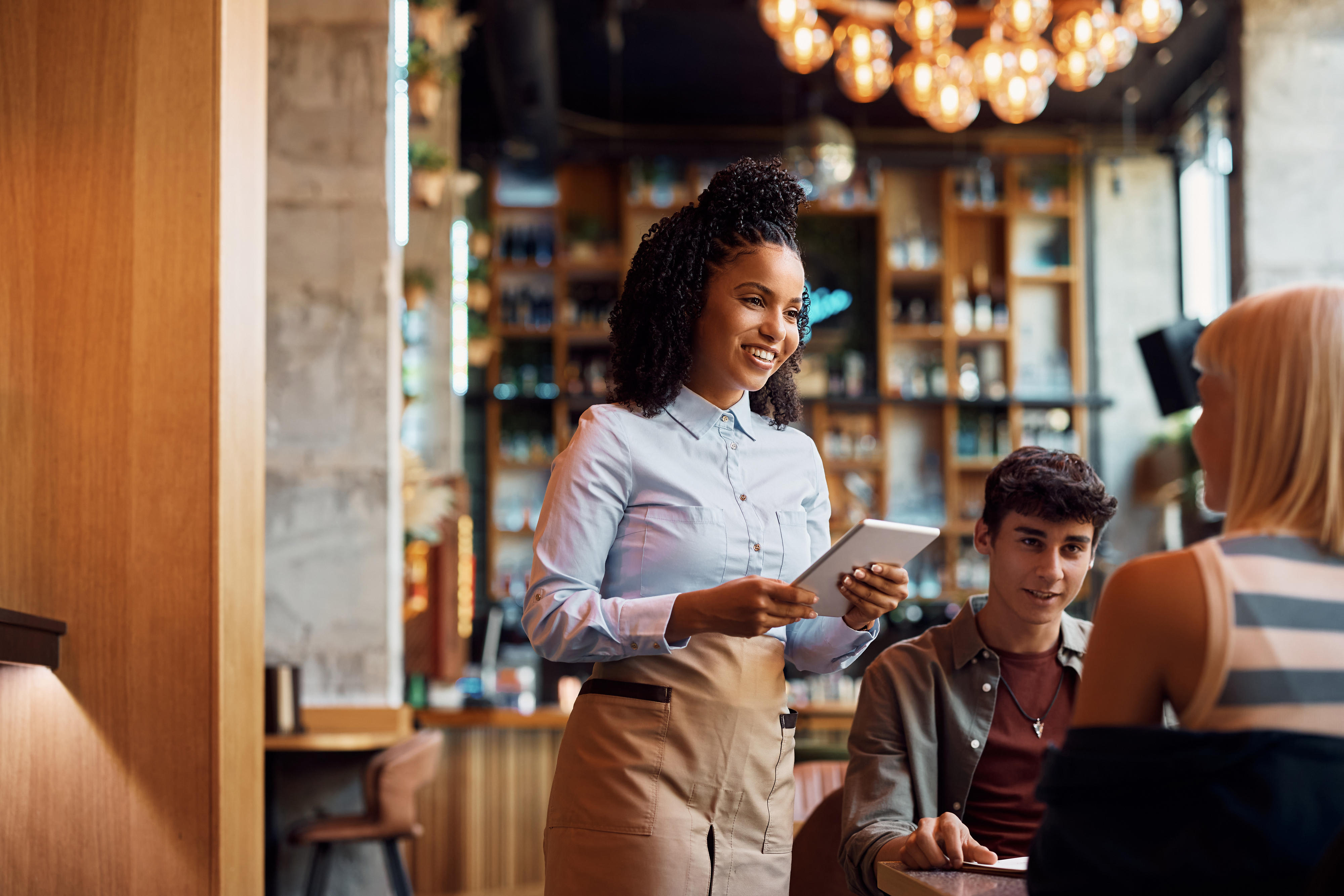 A waitress, holding a digital tablet, takes an order from two customers seated in a modern, well-lit restaurant