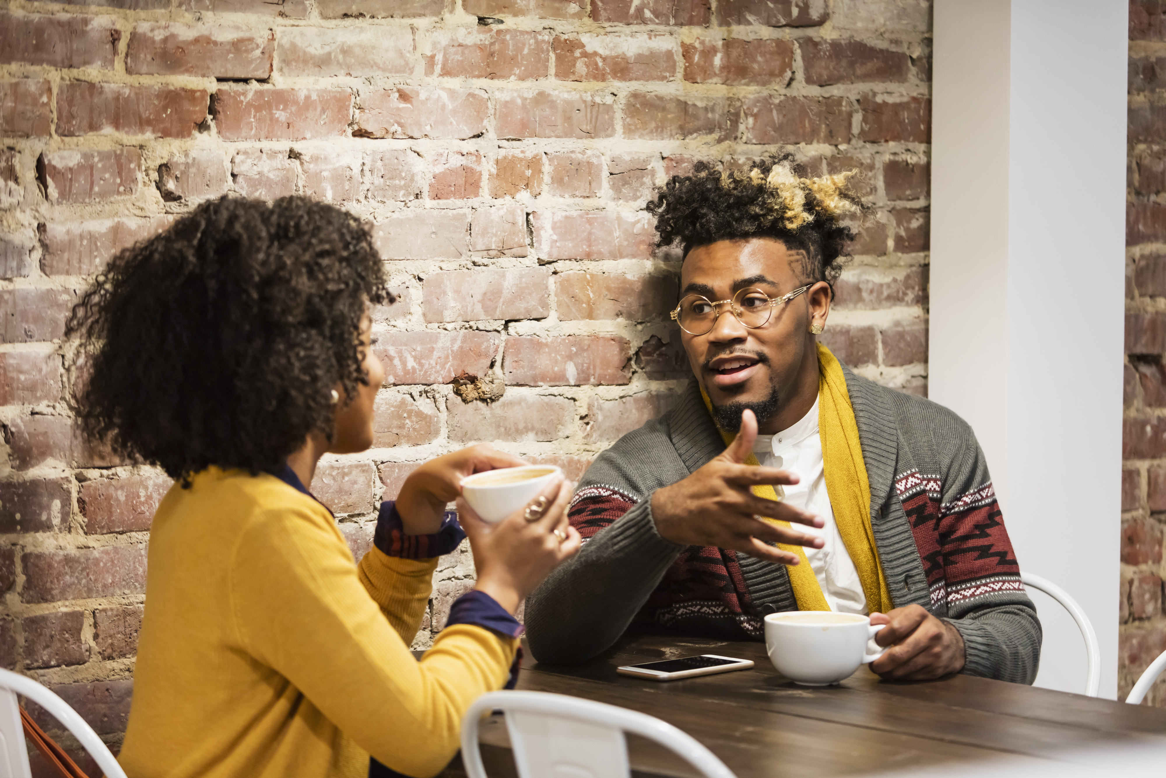Two people having a conversation at a café, holding coffee cups. They are in casual attire, and the setting features a rustic brick wall