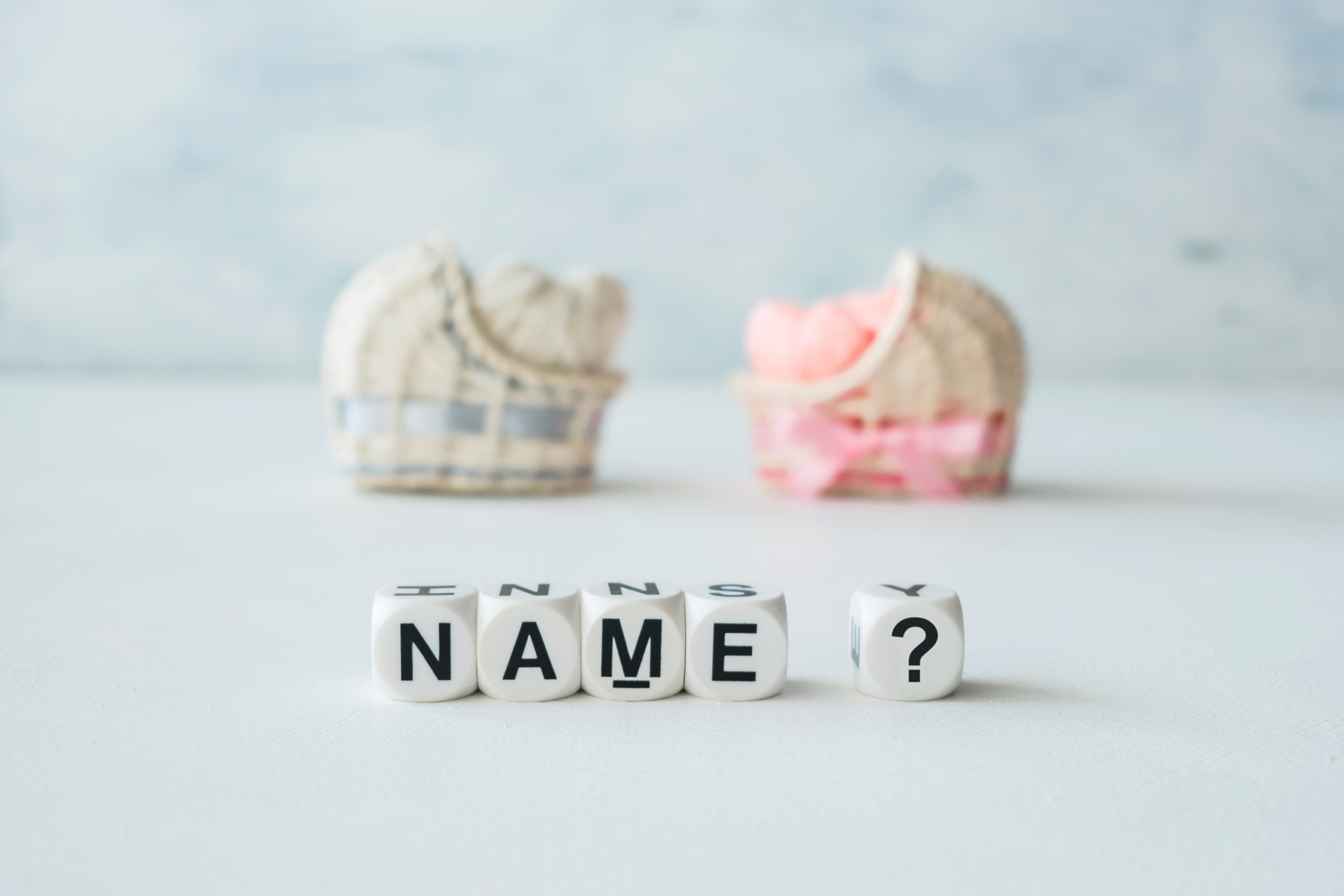 The image shows baby shoes in the background with letter blocks in the foreground spelling out "NAME?". The blocks are in focus, highlighting the question of naming a baby