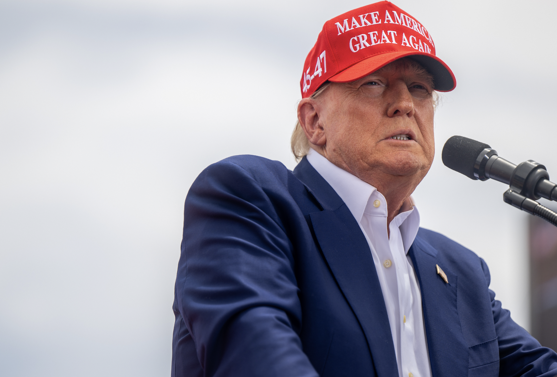 Donald Trump wearing a 'Make America Great Again' hat speaks at a podium outdoors. He is dressed in a suit jacket over a white shirt