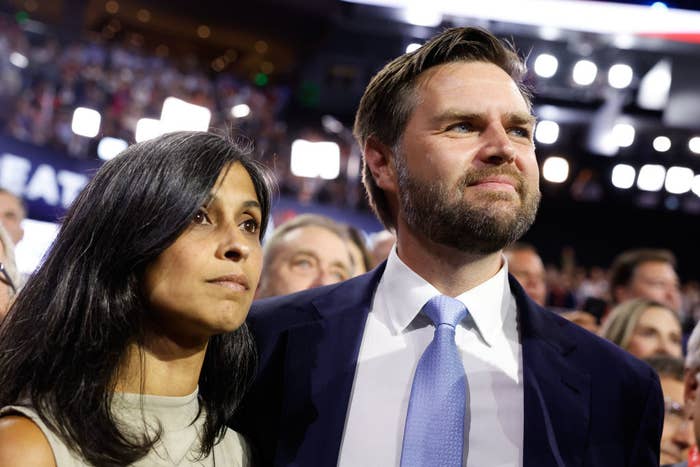 J.D. Vance, wearing a suit and tie, stands with Usha Vance in a sleeveless dress, both looking forward during an event with a crowd in the background