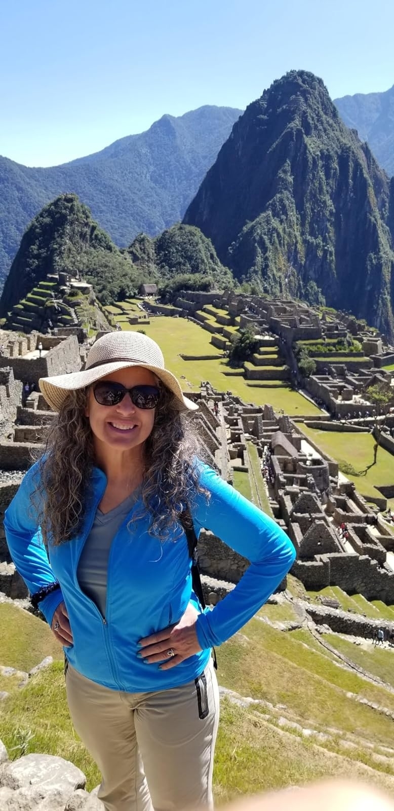 A reviewer with curly hair and a sunhat stands smiling in front of the ancient Inca city of Machu Picchu, with mountains in the background