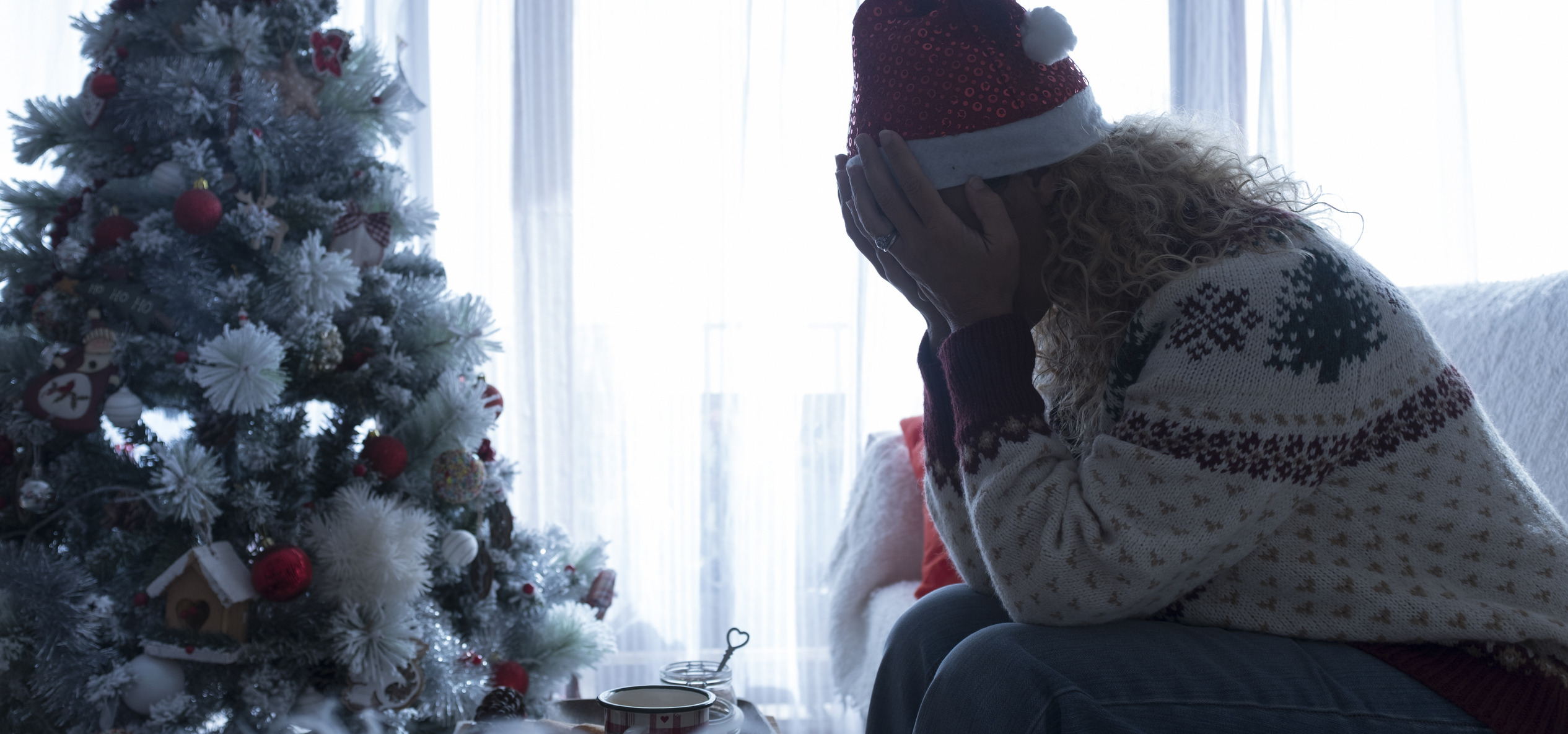 Person in a festive sweater and Santa hat sits with head in hands by a decorated Christmas tree, conveying sadness or stress. Christmas mug nearby