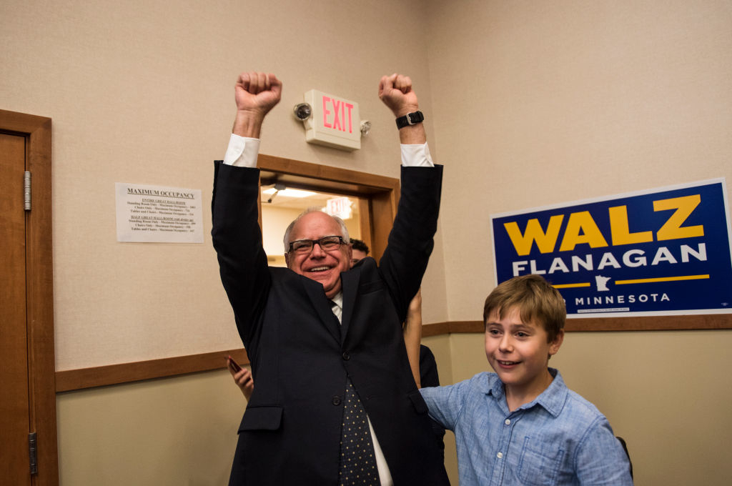 Tim Walz celebrates with his arms raised while a boy stands beside him. A sign reading "Walz Flanagan Minnesota" is visible in the background