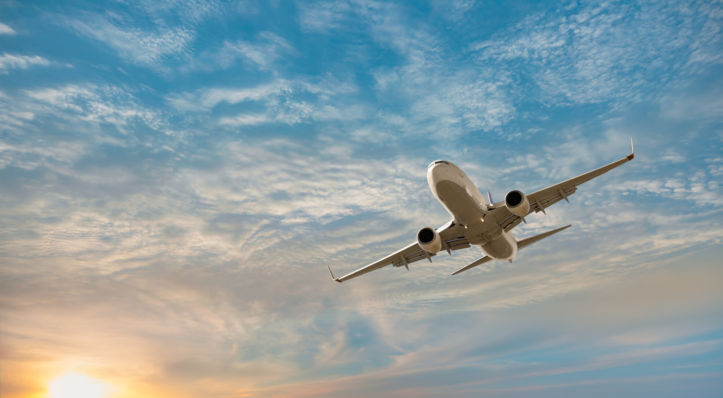 An airplane is flying in the sky with a backdrop of clouds and a glimpse of the sun on the horizon