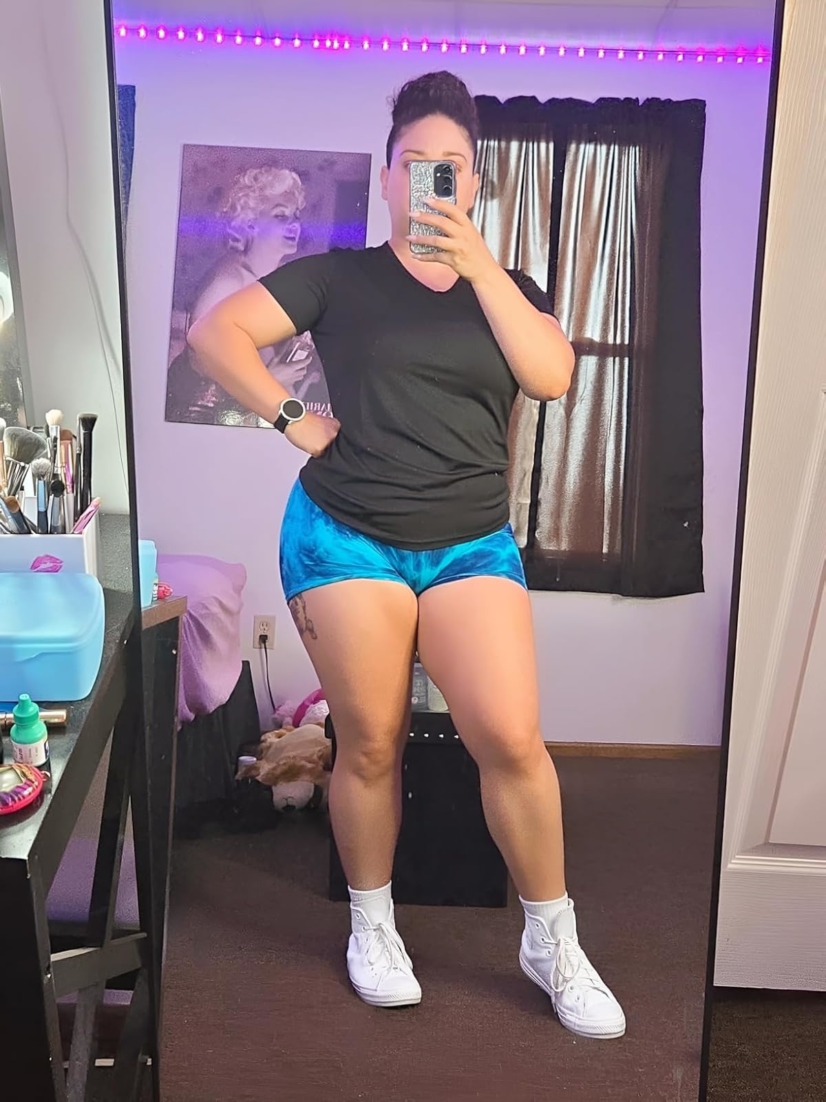 Reviewer's photo of them posing in a bedroom mirror selfie, wearing a black t-shirt, blue athletic shorts, and white sneakers. The room has makeup on a vanity and a poster on the wall
