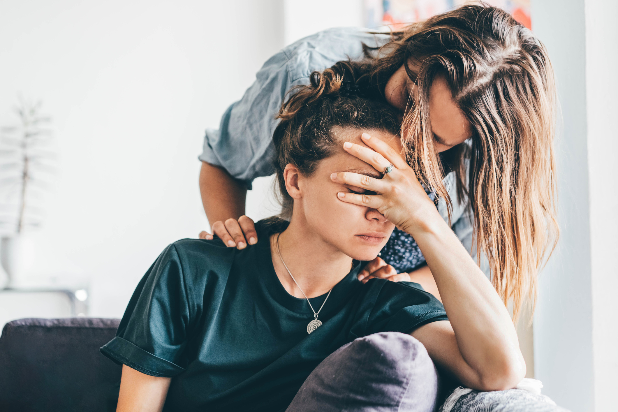 Two individuals share an emotional moment; one person, sitting with their face in their hand, is comforted by another who gently touches their shoulders