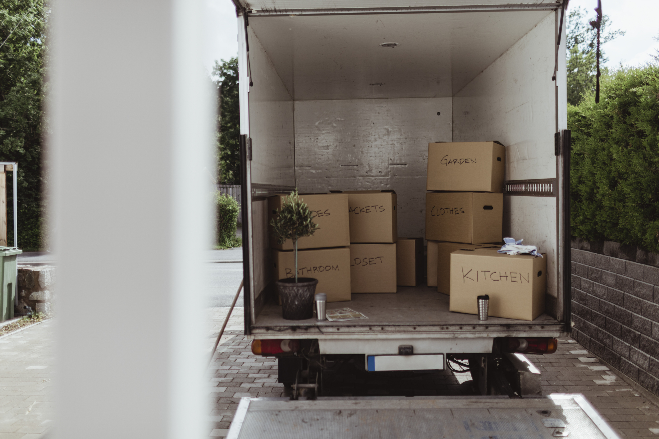 A moving truck is filled with labeled cardboard boxes for bedroom, closet, kitchen, and garden items, and a potted plant sits on the floor of the truck