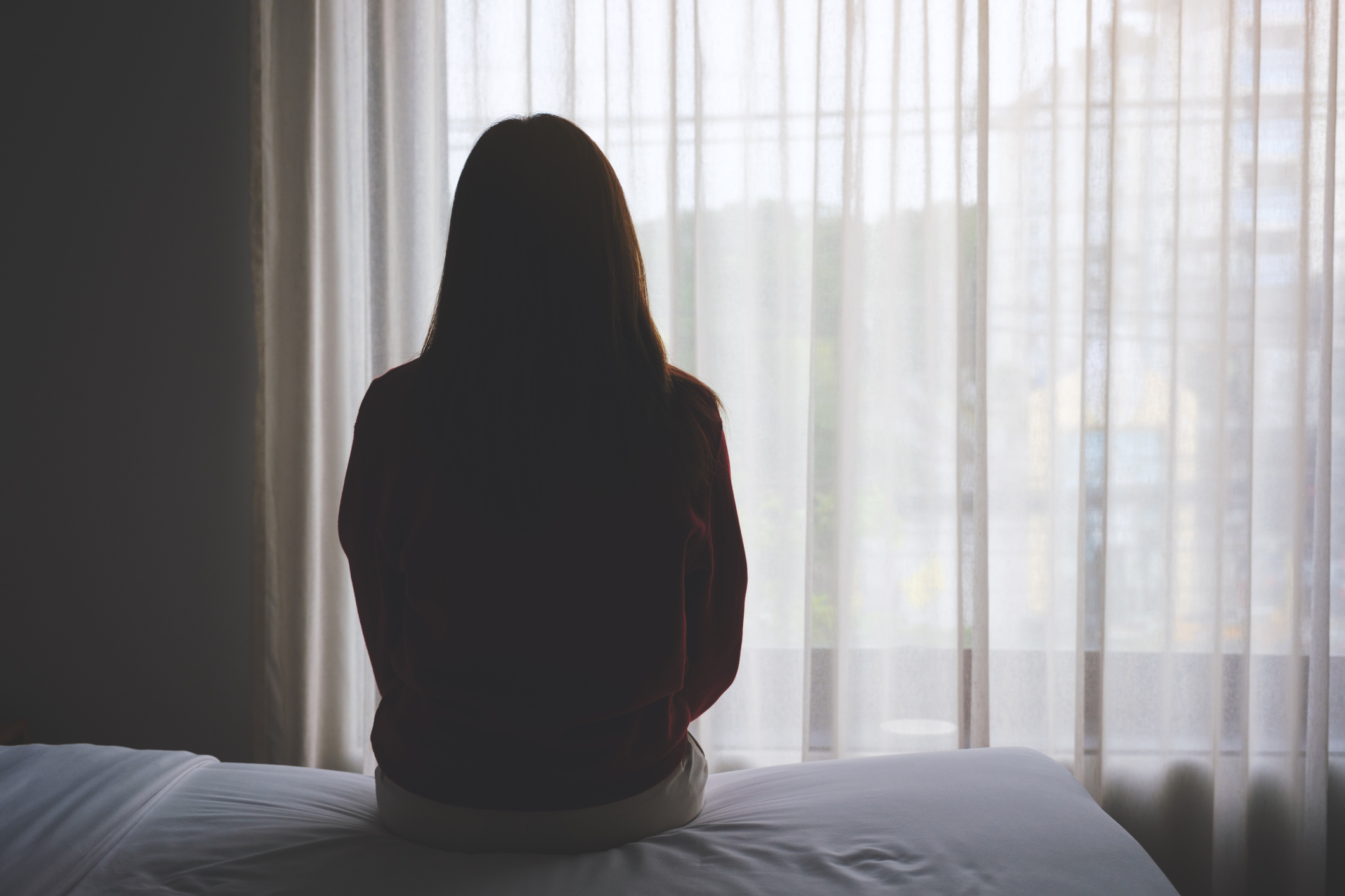A person sits on a bed facing a window with sheer curtains, silhouetted by natural light. The setting suggests contemplation or introspection. No text is present
