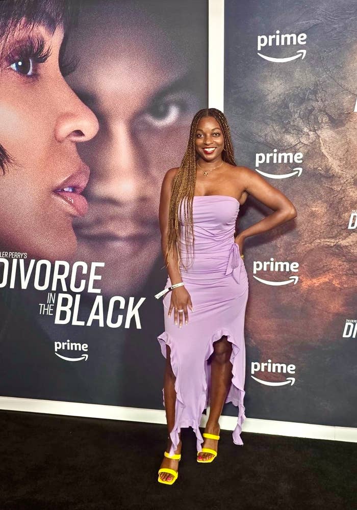 Woman in shoulderless, ruffled dress stands on black carpet at "Divorce in the Black" event with movie poster behind her