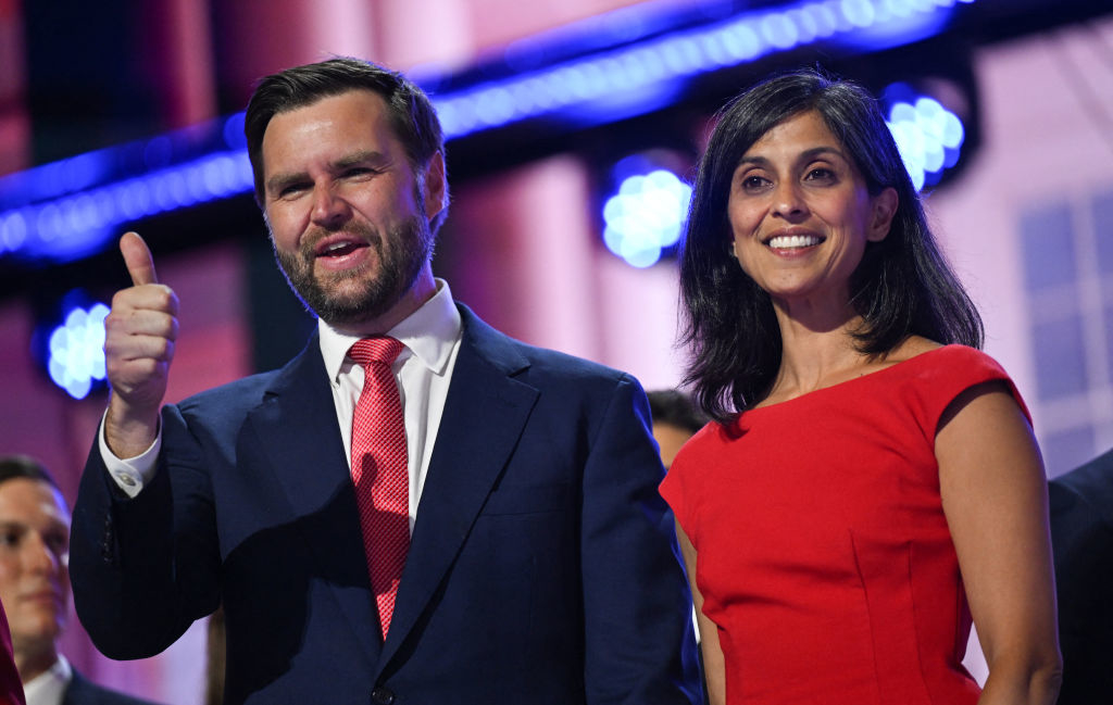 J.D. Vance, in a suit and red tie, gives a thumbs up while standing next to Usha Vance, who is wearing a red dress. Both are smiling