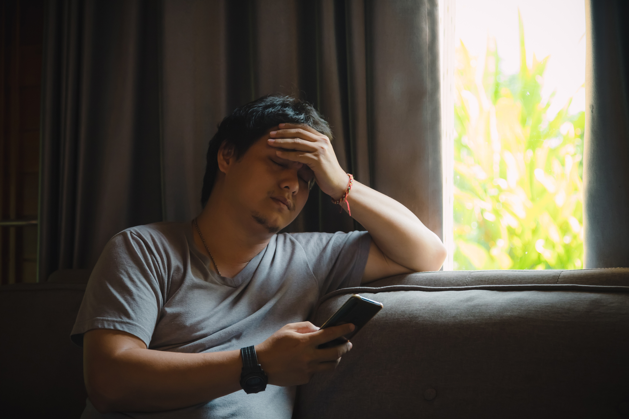 A man sits on a couch with his hand on his forehead and a phone in his other hand, looking stressed near a window