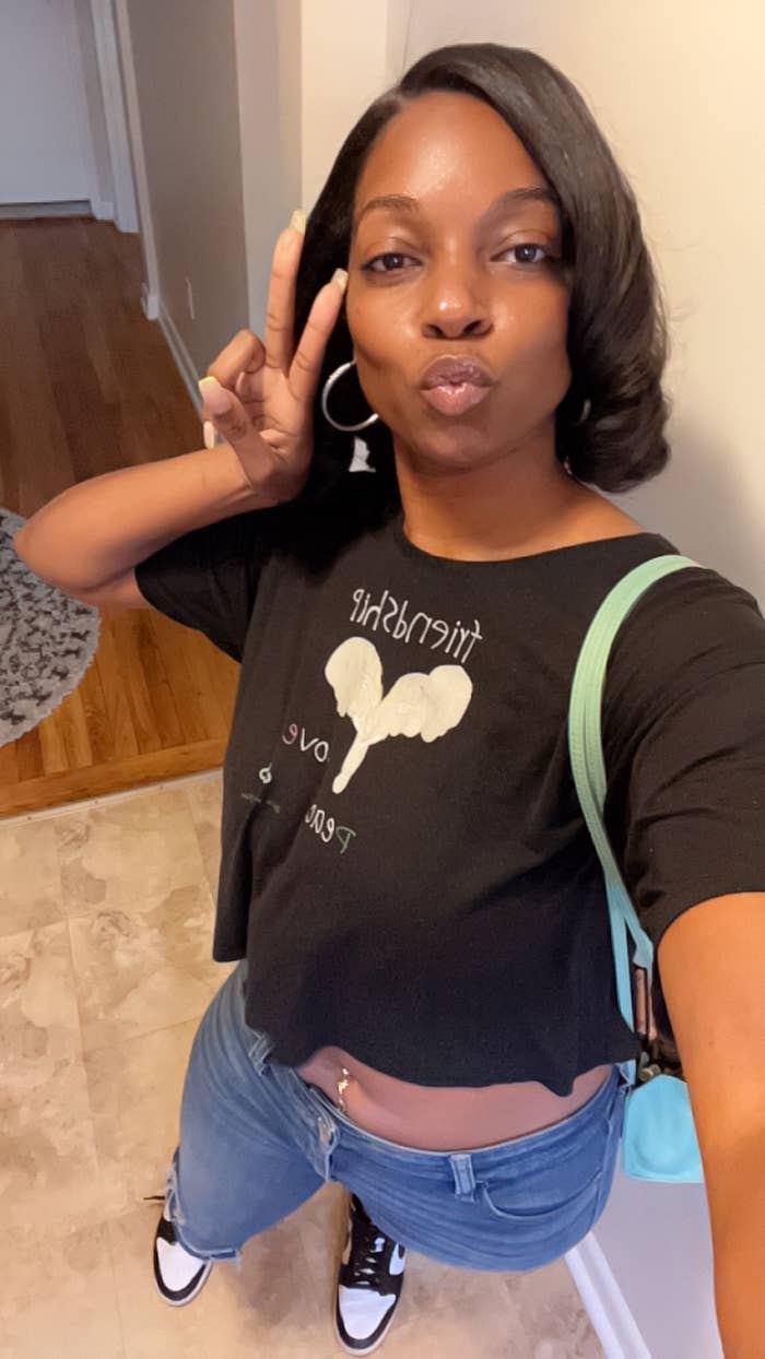 A woman taking a selfie indoors, wearing a cropped T-shirt with text, jeans, and sneakers. She is making a peace sign and pursing her lips