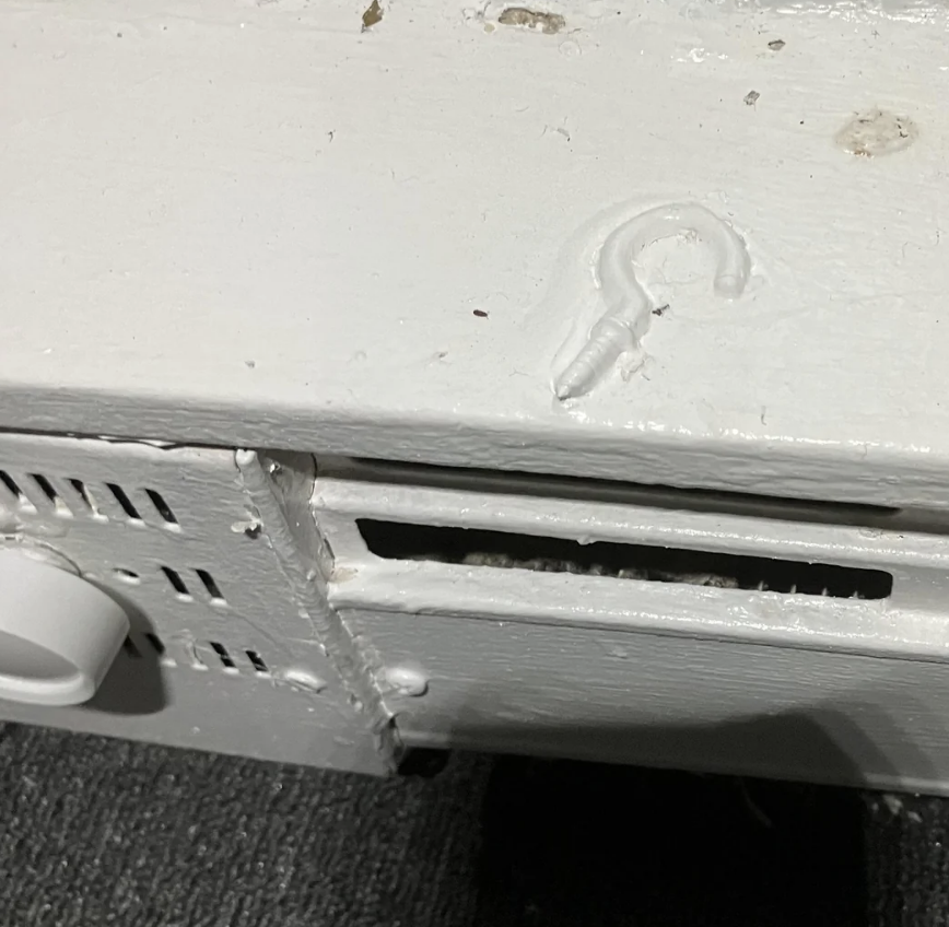 A white-painted metal panel with a partially dented surface, visible ventilation slits, a curved hook-like shape protrusion, and a small open rectangular slot