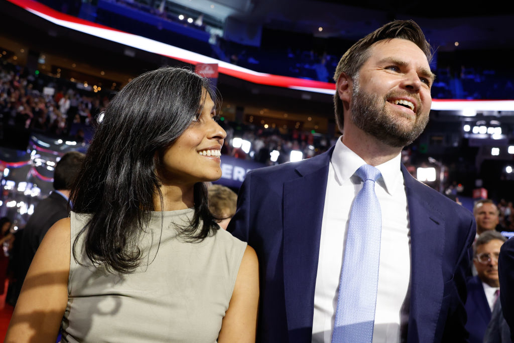 Smiling couple, J.D. Vance in a suit and tie, and Usha Vance in a sleeveless dress, at a public event with a crowd and lights in the background