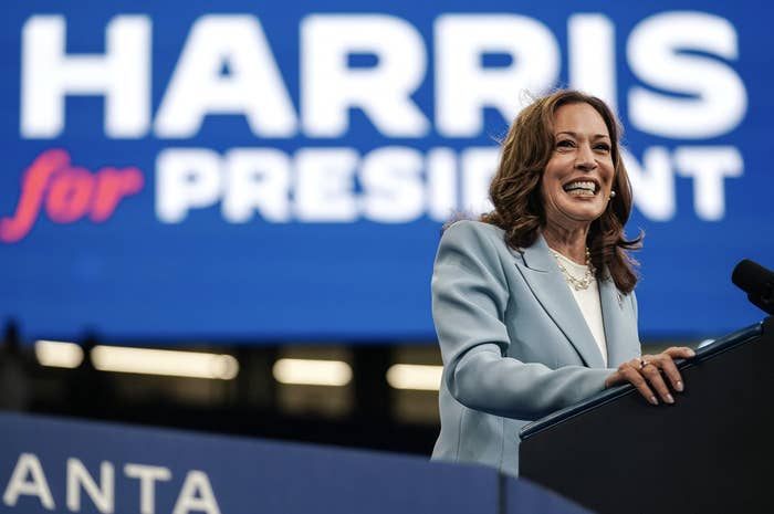 Kamala Harris stands at a podium with a large screen behind her displaying &quot;HARRIS for PRESIDENT.&quot;