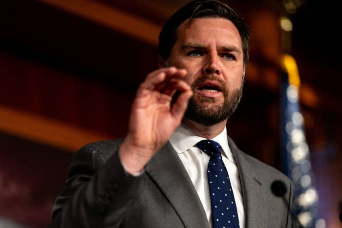 J.D. Vance speaks at a podium, making a hand gesture, with a flag visible in the background. He wears a gray suit and a blue polka dot tie