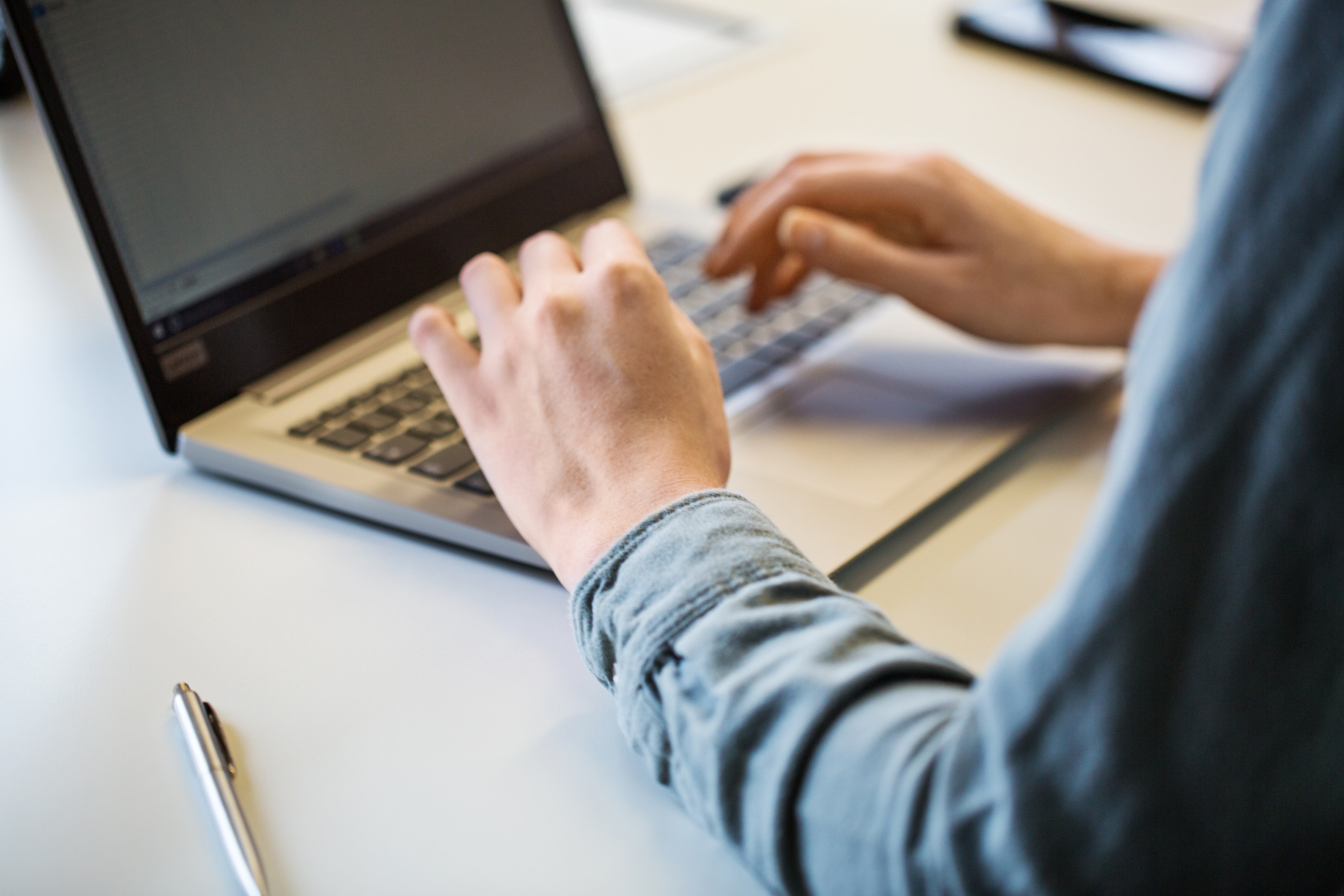 A person types on a laptop at a desk with a pen nearby