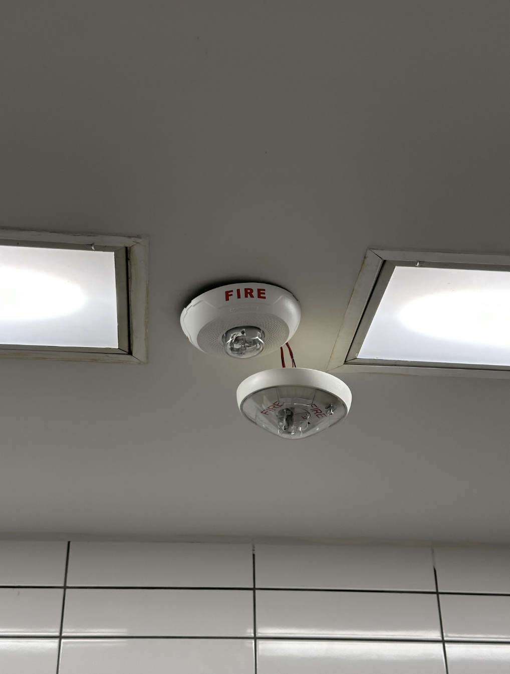 Ceiling-mounted fire alarm and smoke detector near fluorescent lights in a tiled room