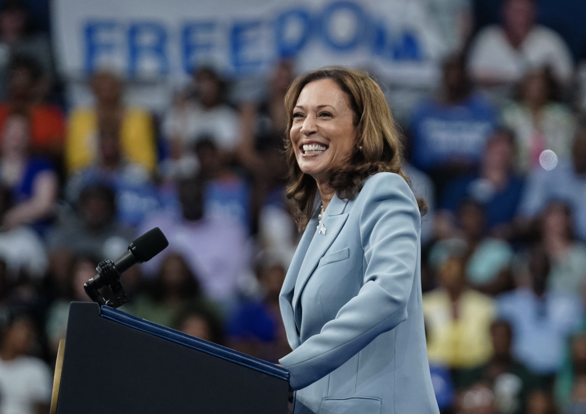 Kamala Harris speaks at a podium during an event with a blurred audience and a &quot;FREEDOM&quot; sign in the background