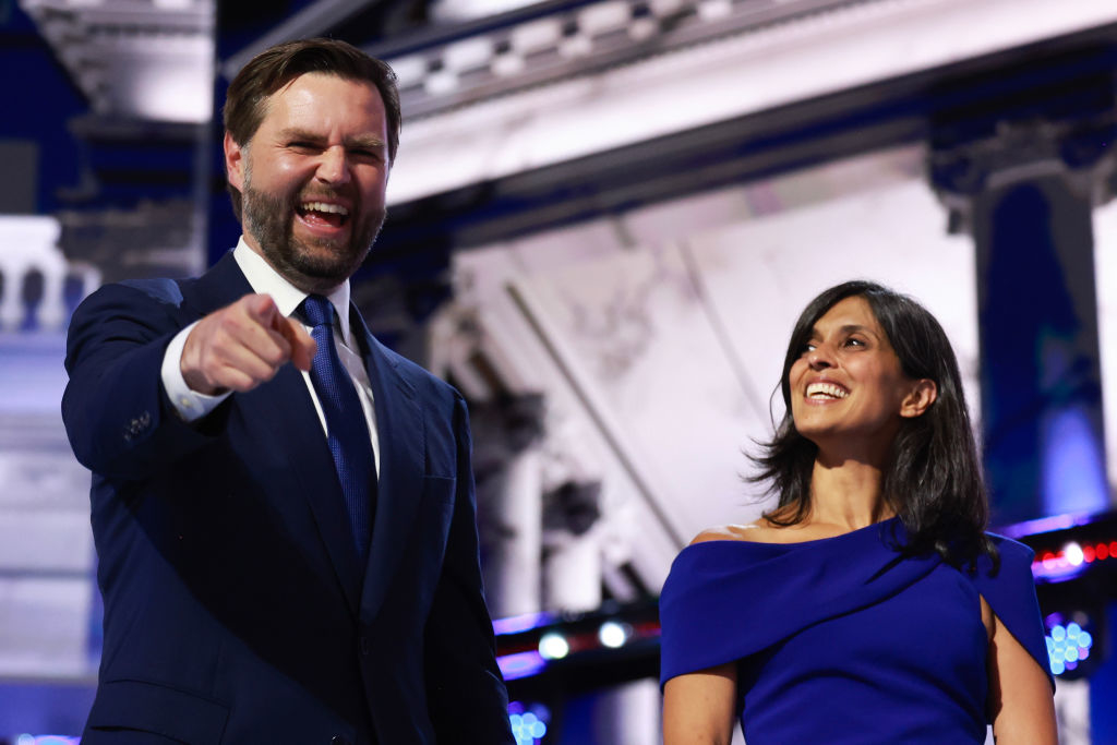J.D. Vance, in a suit, points with his right hand while smiling, and Usha Vance, in a one-shoulder dress, stands beside him, both on a stage
