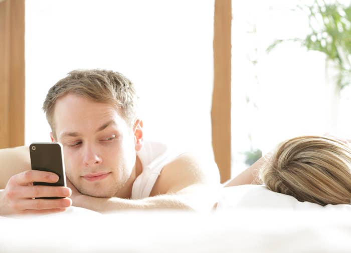 A man looks at a smartphone over his shoulder while lying next to a woman sleeping in bed