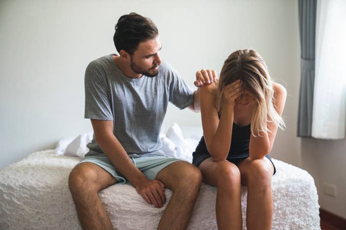 A man comforts a woman sitting on a bed with her head in her hands. Both appear distressed. The man is in casual wear; the woman is in a sleeveless dress