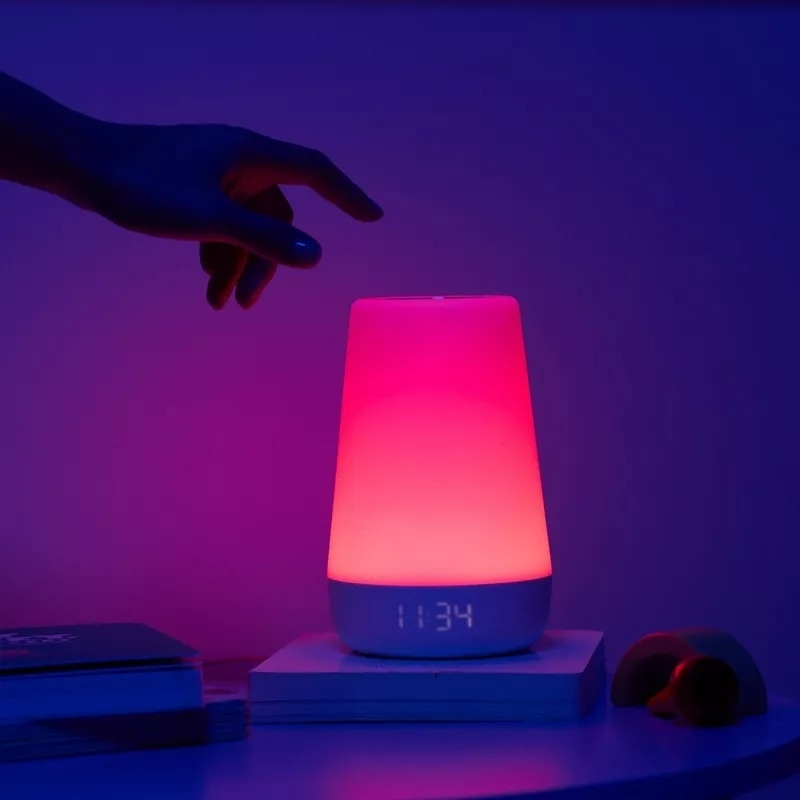 A hand reaches towards a glowing smart lamp with the time 11:34 displayed on it, sitting on a bedside table next to books