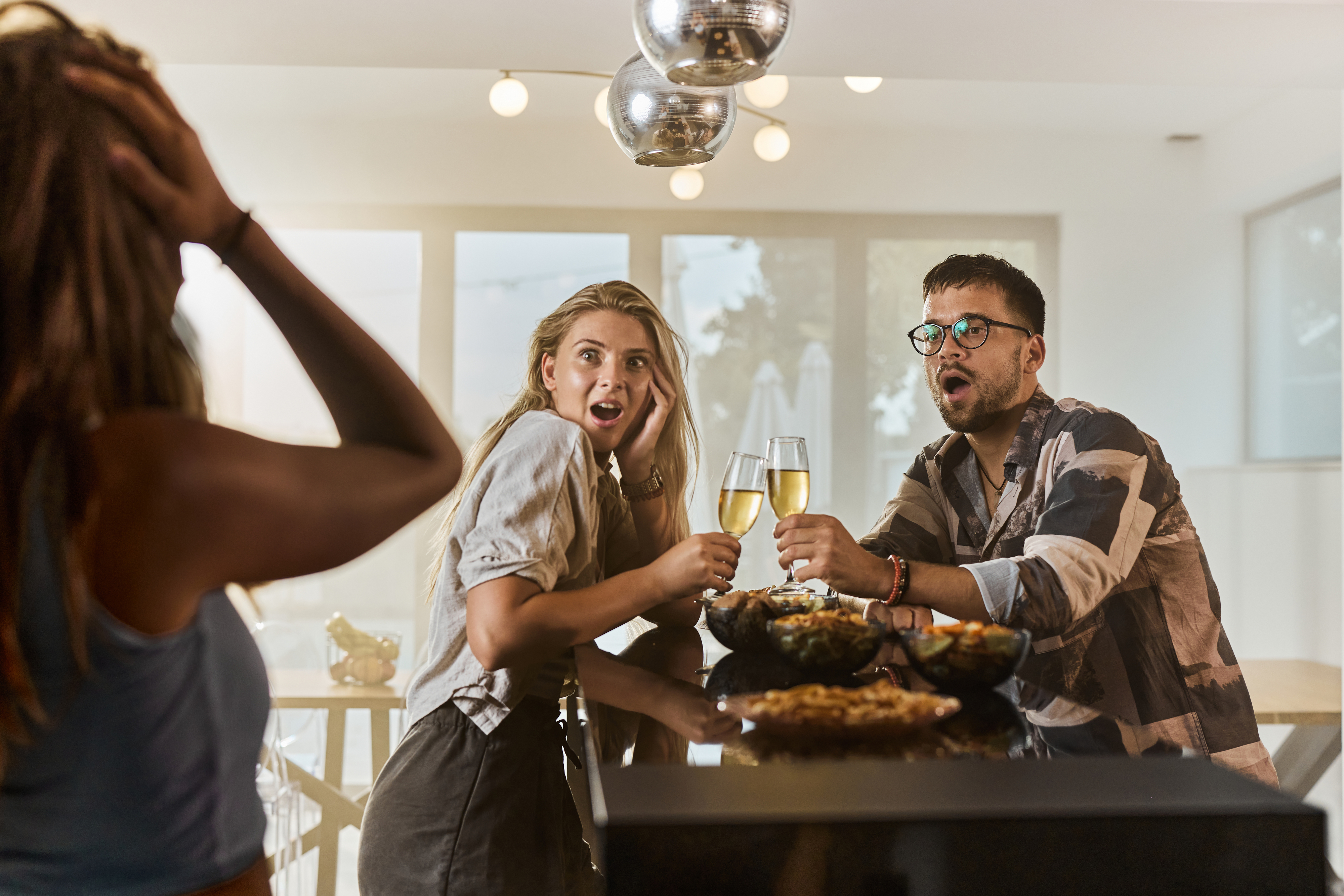 Three people reacting in surprise around a table with food and drinks, two holding glasses of champagne