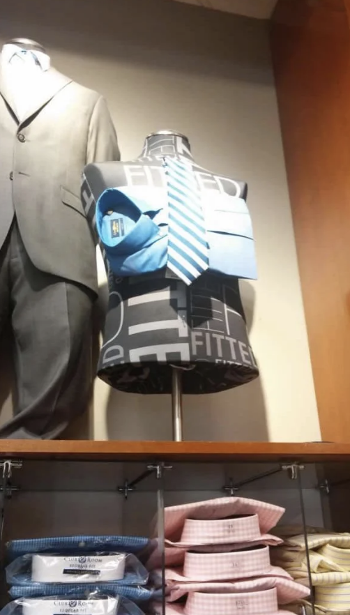 Mannequin displaying a blue-striped tie with a blue shirt, folded pants, and a jacket on the upper half. Gray suit on another mannequin next to it. Shirt packages below