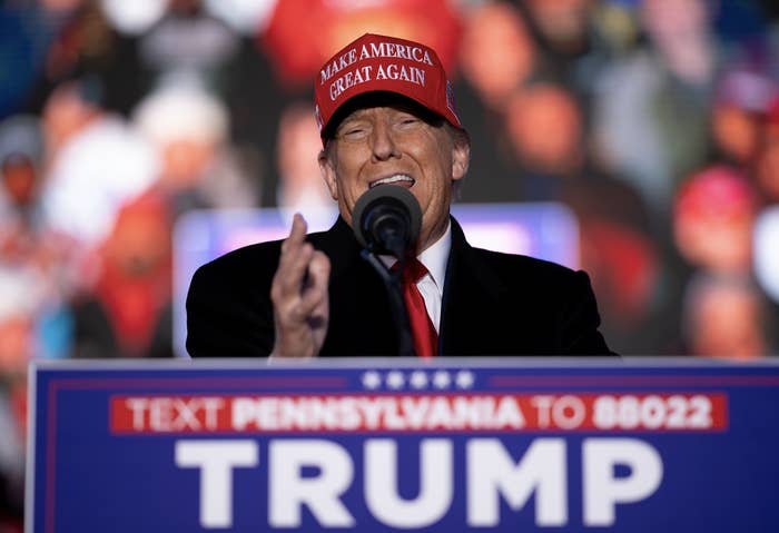 Donald Trump speaking at a rally. He is wearing a red &quot;Make America Great Again&quot; hat and a black suit with a red tie. The podium has a sign with text, &quot;TEXT PENNSYLVANIA TO 88022 TRUMP.&quot;
