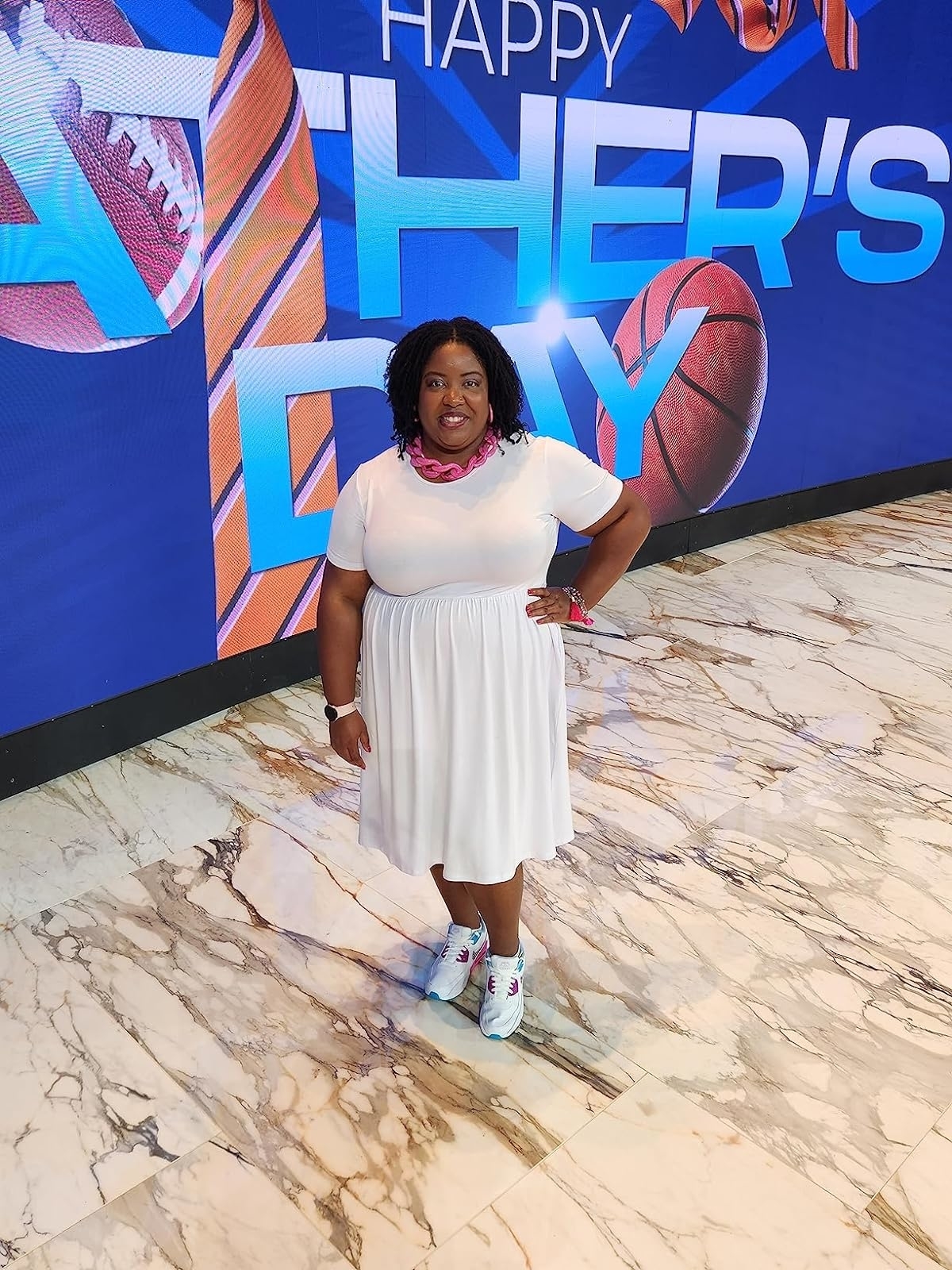 Reviewer's photo of them standing in front of a large "Happy Father's Day" sign, wearing the dress in the color white, styled with a chunky pink necklace and white sneakers
