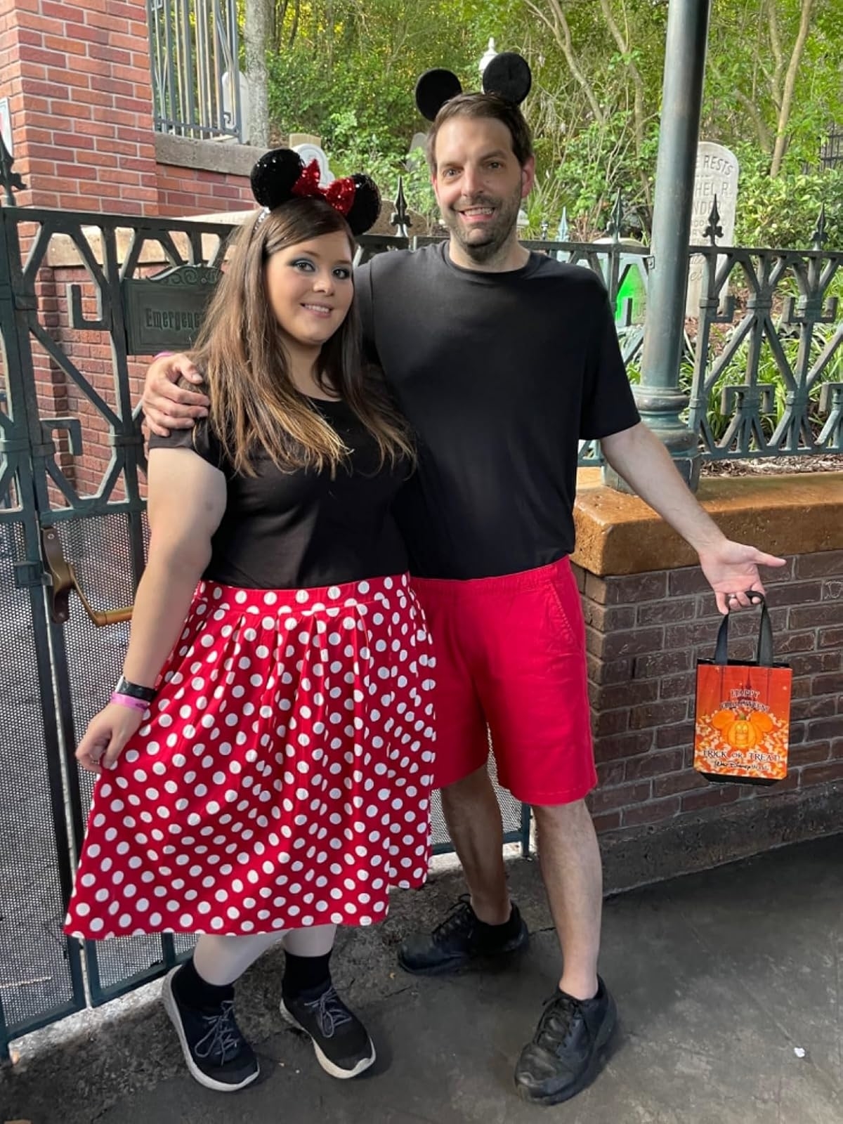 Reviewer's photo of two adults, both wearing Mickey and Minnie Mouse-themed clothing and ears, pose outdoors with a Halloween-themed gift bag