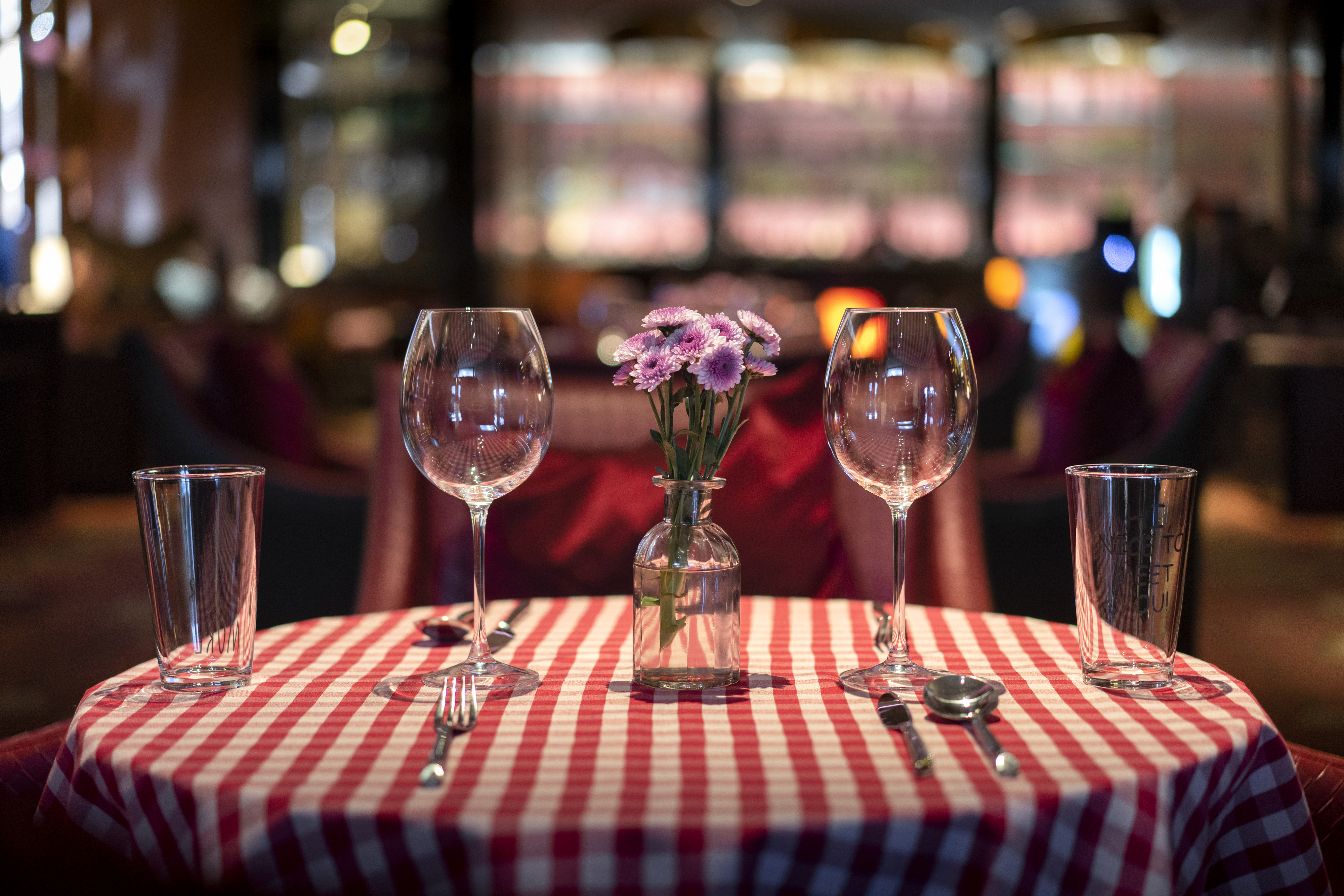 A table with red and white checkered tablecloth set with two wine glasses, two water glasses, and silverware, adorned with a small vase of purple flowers
