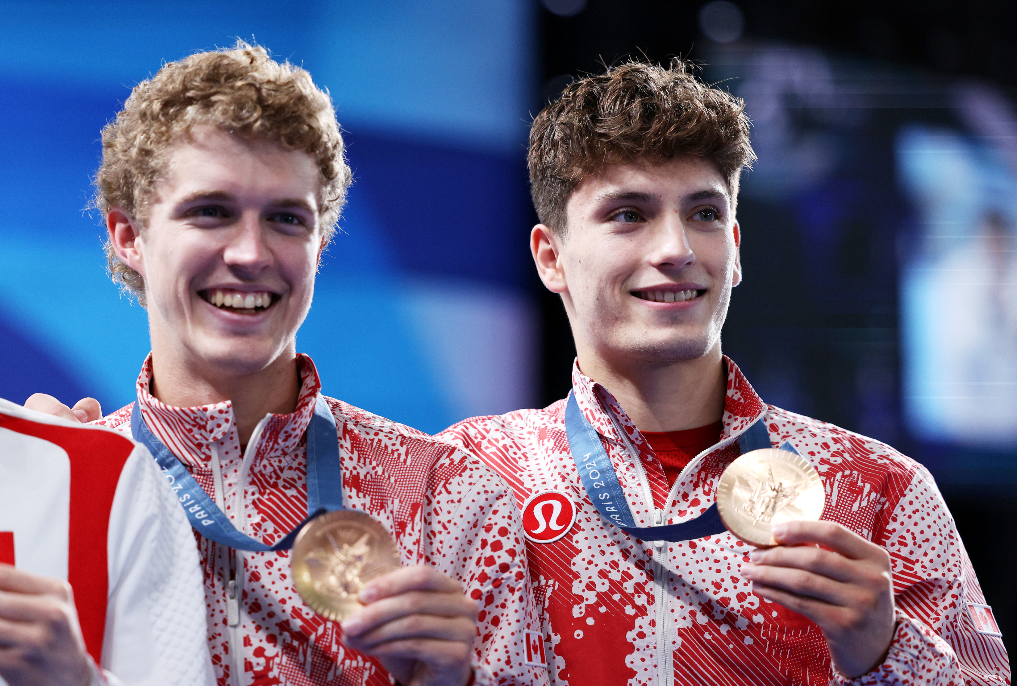 Two athletes smile while holding medals on a podium, wearing matching sports jackets with athletic branding