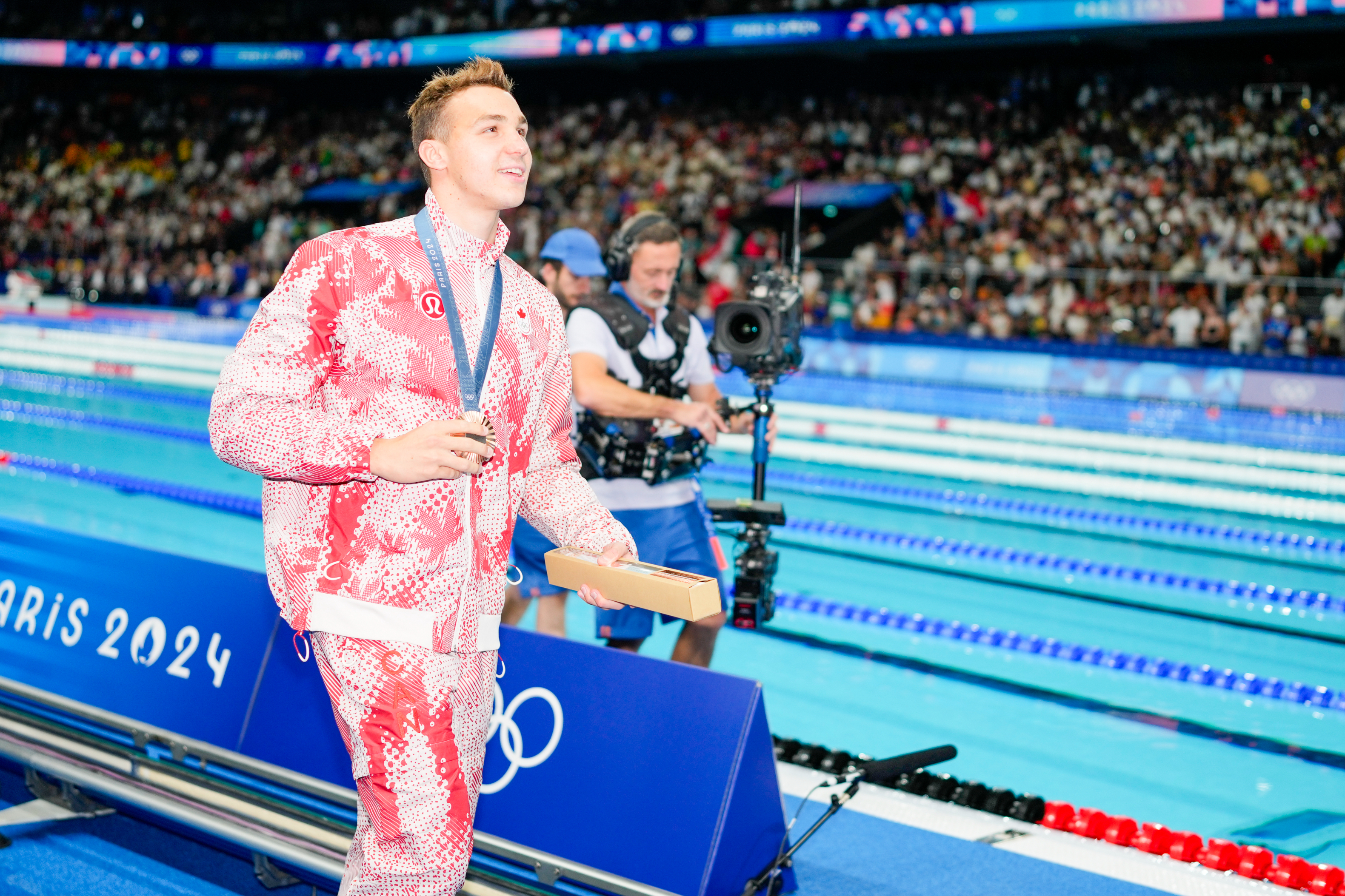 Olympic swimmer dressed in patterned sportswear with a medal around his neck, walking poolside at Paris 2024 event with spectators and media in the background