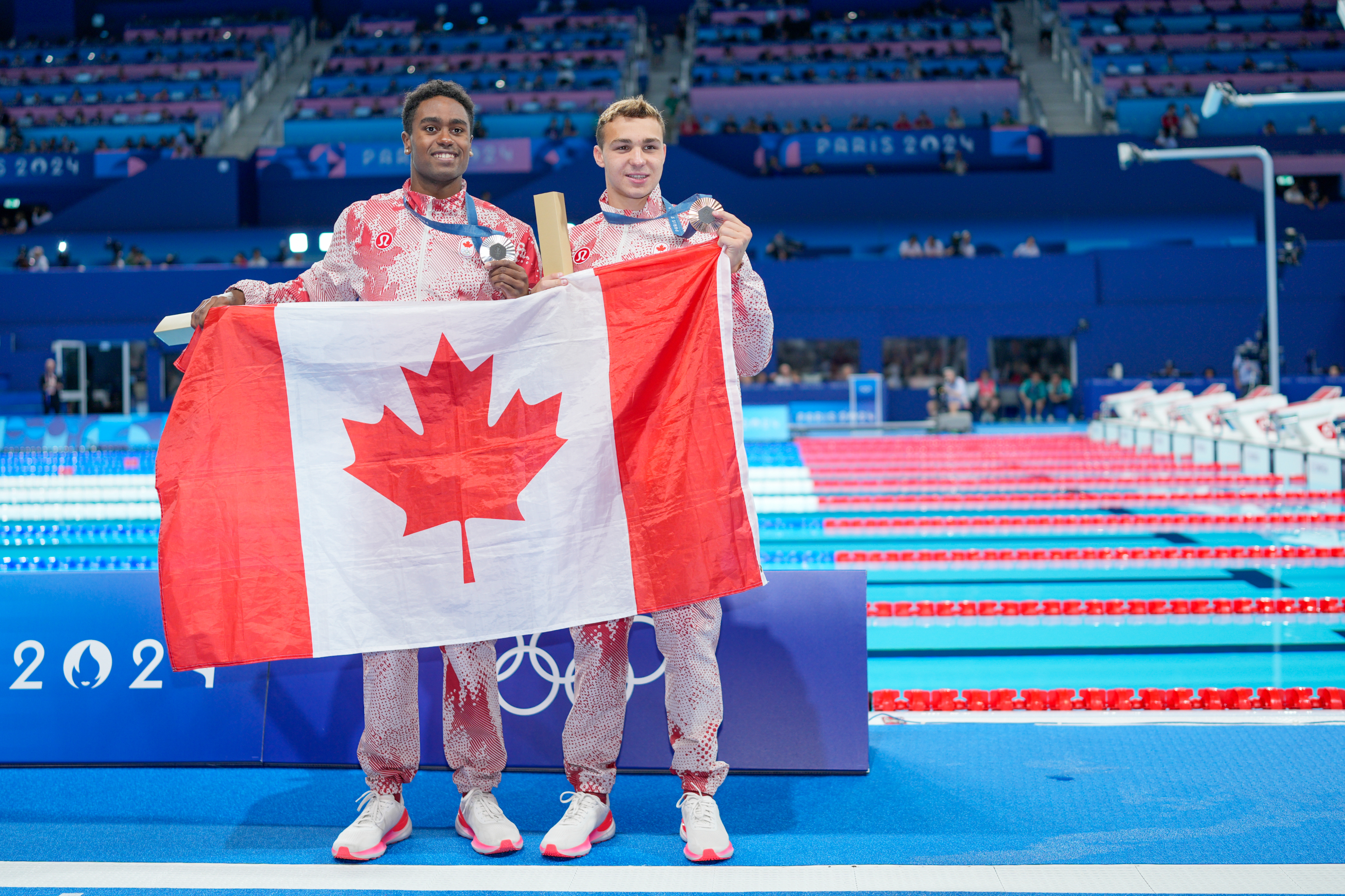 Canadian swimmers stand on the podium holding a flag after winning a medal at the 2024 Olympics. They wear matching athletic gear with maple leaf patterns