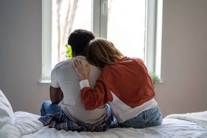 Two people sit on a bed, with one person's arm wrapped around the other in an affectionate manner. Their faces are not visible