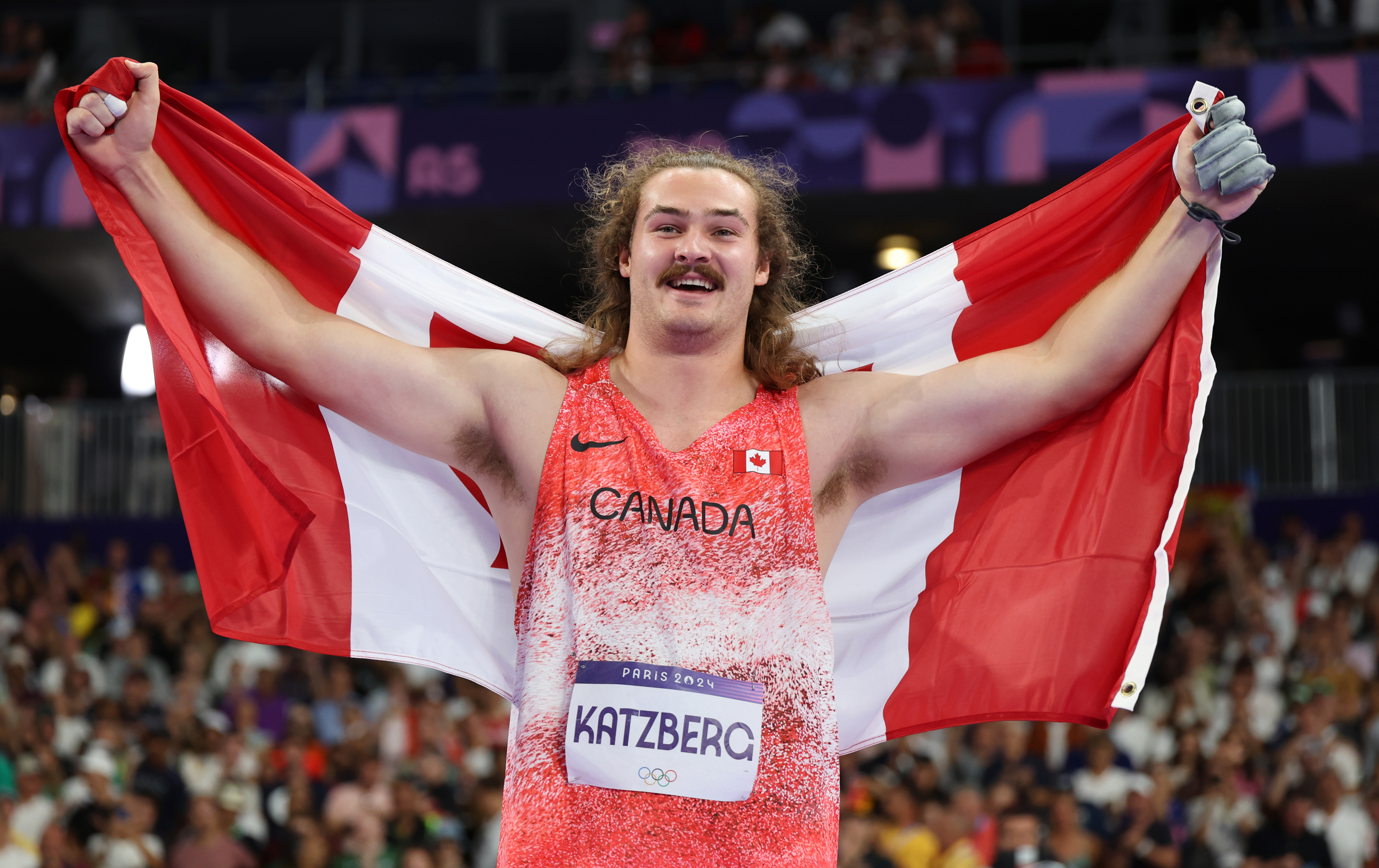 Cameron Katzberg stands triumphantly on a sports field, holding the Canadian flag behind him, wearing a "Canada" jersey during the Paris 2024 Olympics