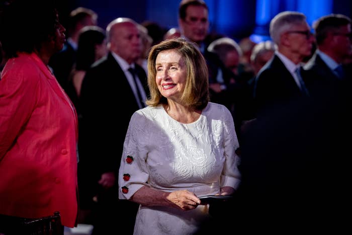 Nancy Pelosi stands smiling in a crowd at an event, wearing an elegant white dress with rose details on the sleeves