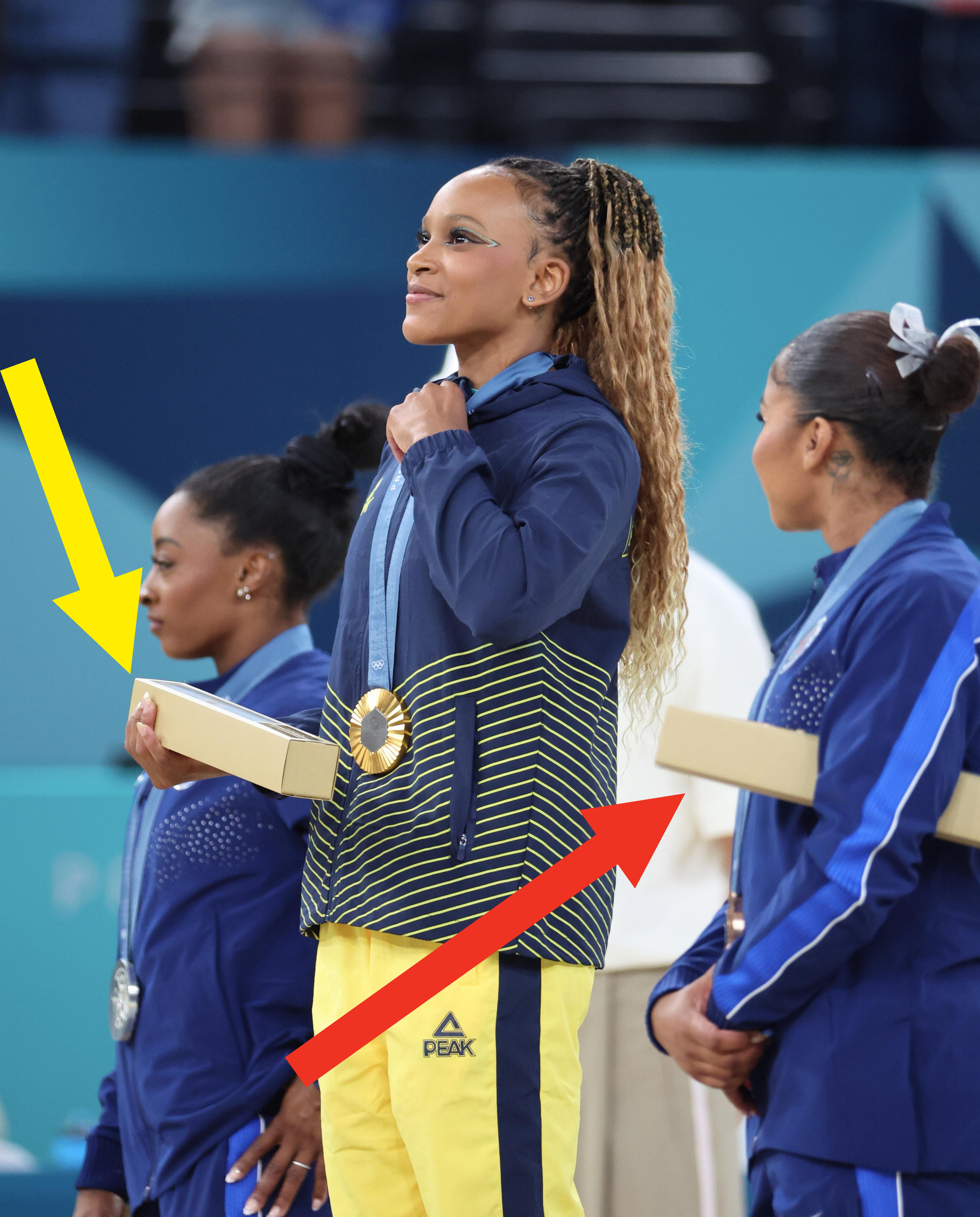 Simone Biles, Sunisa Lee, and Jordan Chiles on the awards podium wearing their medals and team tracksuits, holding gift boxes