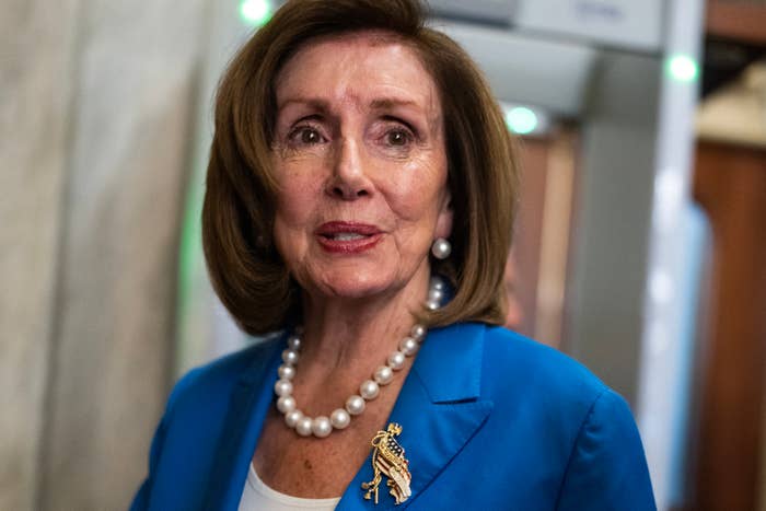 Nancy Pelosi, wearing a blue blazer and pearl necklace, stands in front of a background that includes a doorway