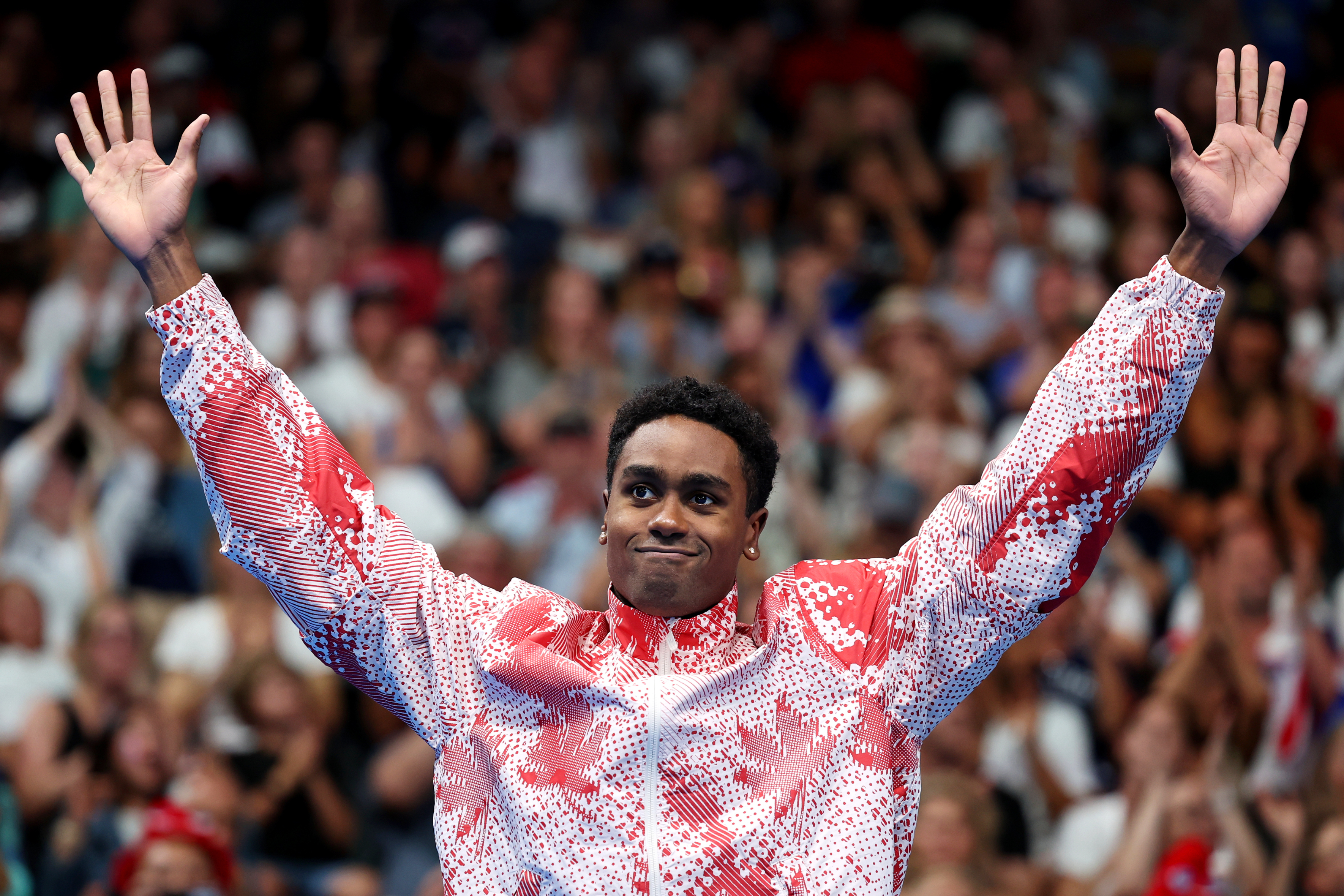 Track athlete Noah Lyles raises his arms in victory, wearing a patterned jacket, in front of a cheering crowd