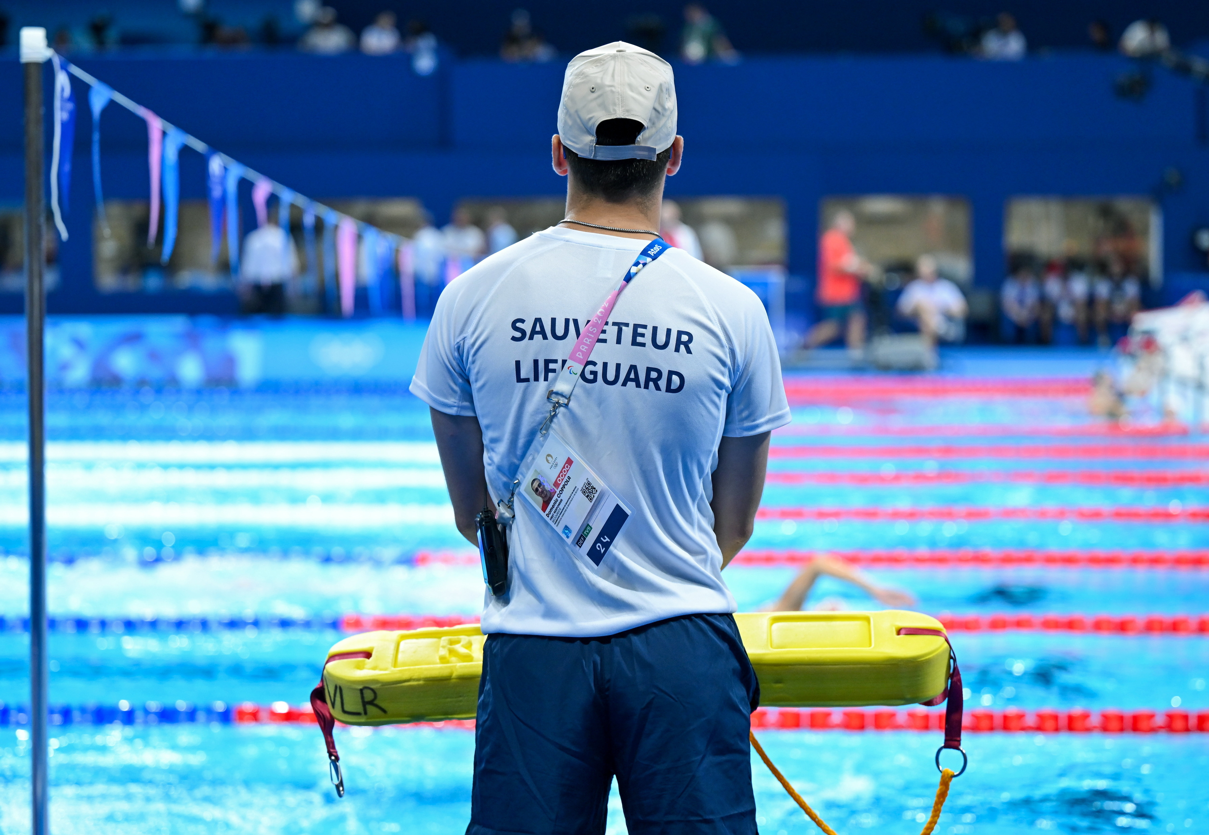 A lifeguard wearing a white t-shirt and a cap stands poolside with a rescue float, overseeing a swimming competition. Text on the shirt reads "SAUVETEUR LIFEGUARD."