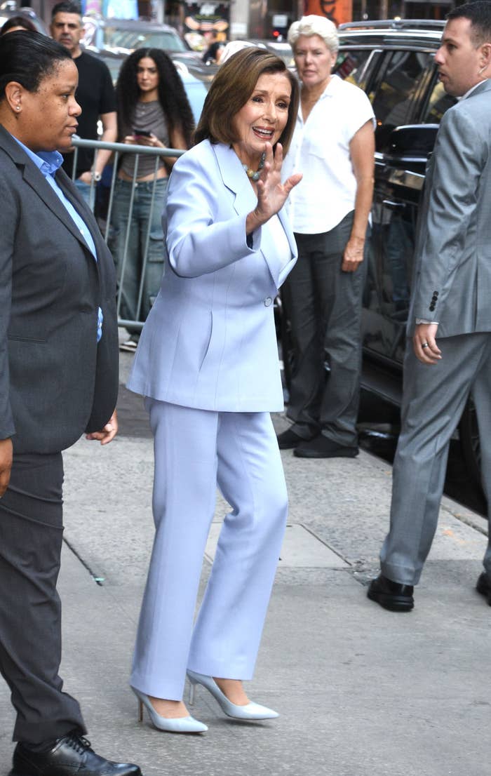 Nancy Pelosi waves while walking, dressed in a stylish suit. She is accompanied by security personnel and surrounded by other people