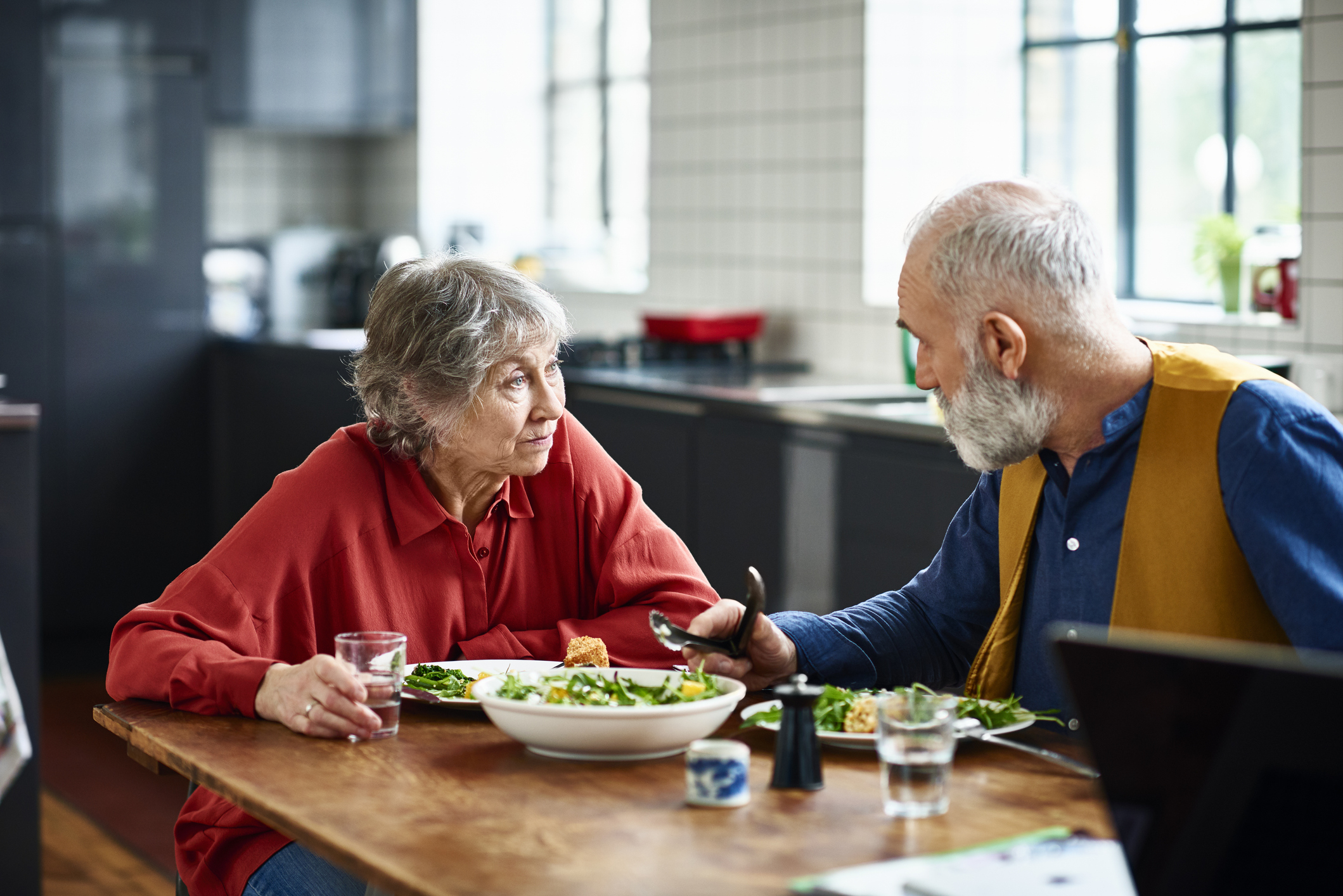 An elderly woman and man are sitting at a kitchen table eating a meal, engaged in conversation. The man is wearing a yellow vest over a blue shirt; the woman is in a red top
