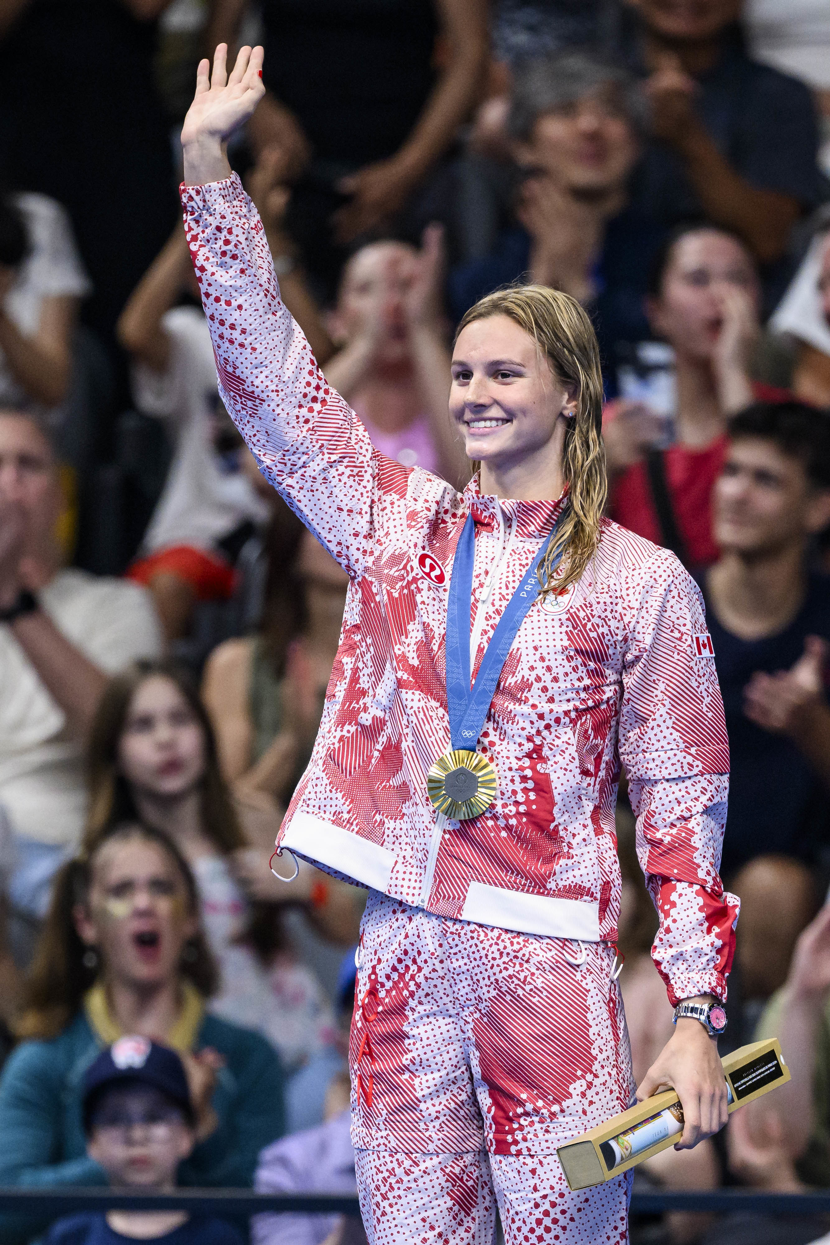 Swimmer standing on a podium, smiling and waving while wearing a tracksuit and medal, holding an award. Crowd clapping and cheering in the background